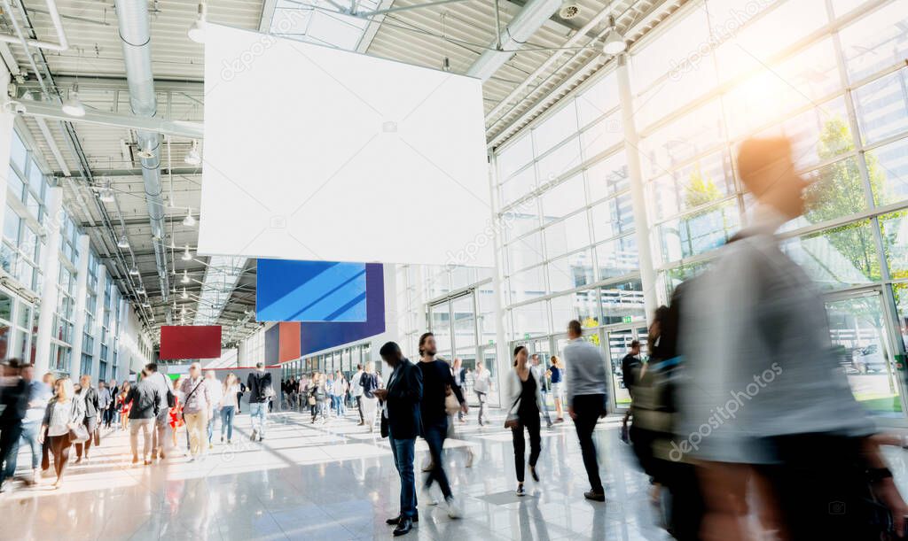 Many trade fair visitors walk under a blank billboard through a hall