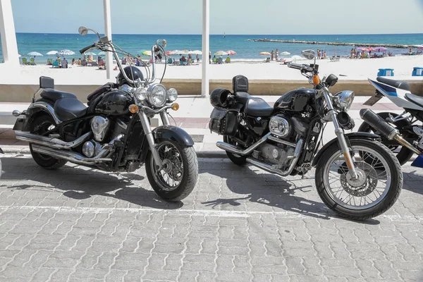 horizontal view of two motorbikes parked on the street with the beach in the background