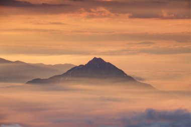 Conical Storzic peak emerges from sea of clouds and translucent morning mist with cloud stripes in the sky illuminated by red sunlight at autumn sunrise, Kamnik Savinja Alps Carniola Slovenia Europe clipart