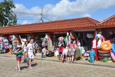 Anapa, Rusya, 18 Temmuz 2018. Anapa'daki gezinti yolunda plaj aksesuarları, giysiler ve Hediyelik Eşyalar ile küçük pavyonlar yakınında yürüyen insanlar