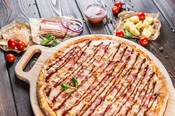 Macro. Meat pizza with red barbecue sauce, bacon and chicken on a round cutting board on a dark wooden background. Igredients