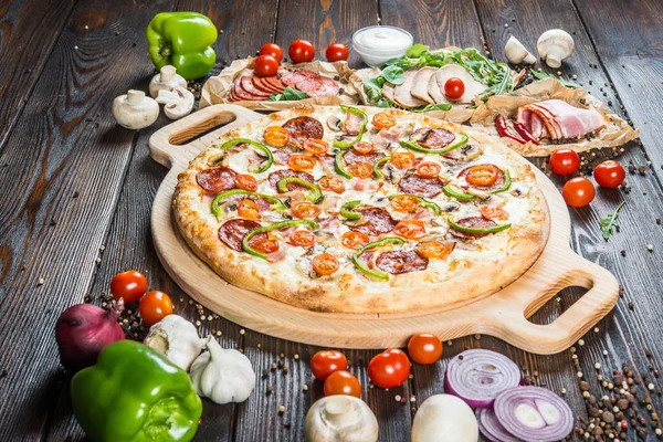 Large meat pizza with salami, bacon, mushrooms, tomatoes and paprika on a round cutting board on a dark wooden background. Ingredients.