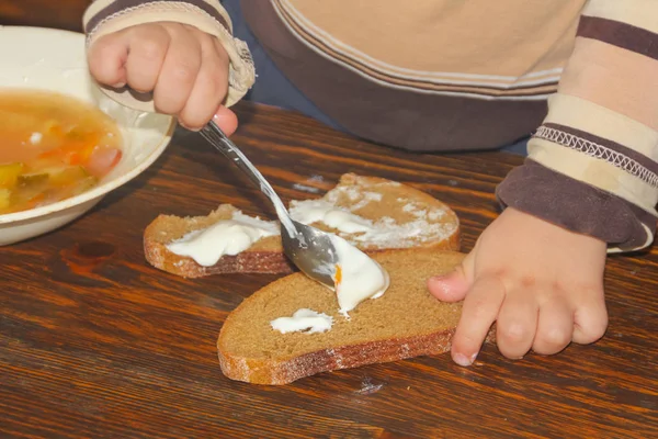 Un niño unta crema agria con pan. untar la crema en un rollo . — Foto de Stock