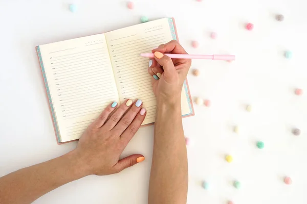 Γυναικεία χέρια με στυλό στο σημειωματάριο και πολύχρωμες μπάλες στο τραπέζι. Κομψό μοντέρνο χρώμα γυναικείο μανικιούρ — Φωτογραφία Αρχείου