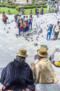 Melon şapkalı iki Bolivyalı kadın La Paz 'daki Merkez Meydanı' ndaki Plaza Murillo 'da oturuyor ve arkada La Paz Bolivya Katedrali var.