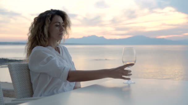 Una giovane donna seduta sulla spiaggia incontra un tramonto con un bicchiere di vino in mano — Video Stock