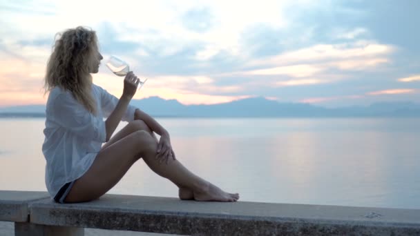 Una giovane donna seduta sulla spiaggia incontra un tramonto con un bicchiere di vino in mano — Video Stock