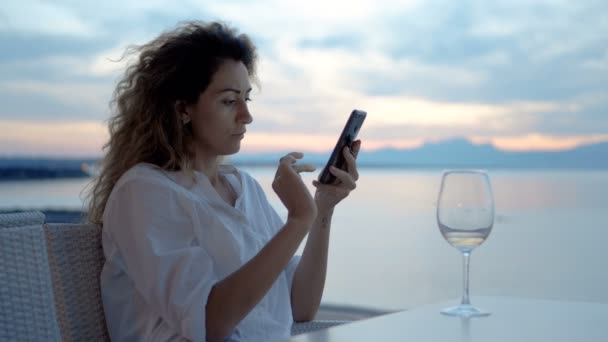 Una mujer joven toma una foto de una puesta de sol y una copa de vino en el teléfono en la costa del océano — Vídeo de stock