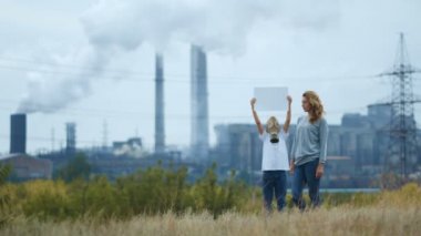 Anne ve oğlu gaz maskeli bir fabrika ve lüle arka planda dur. Elinde bir işaret tutan