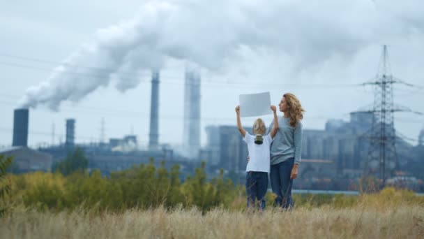 Mamá e hijo en una máscara de gas de pie en el fondo de una fábrica y pipas de fumar. En las manos sosteniendo un cartel — Vídeos de Stock