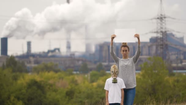 Mãe e filho em um carrinho de máscara de gás no fundo de uma fábrica e cachimbos de fumar. Nas mãos segurando um sinal — Vídeo de Stock