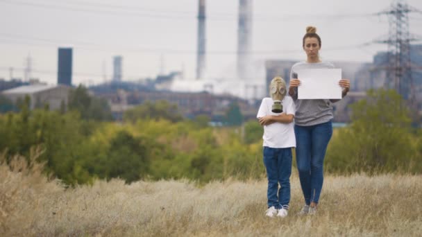 Máma a syn v plynové masce stojí v pozadí továrny a dýmky. V rukou drží ceduli — Stock video