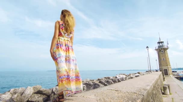 Una chica en un vestido está de pie en el océano mirando el faro — Vídeos de Stock