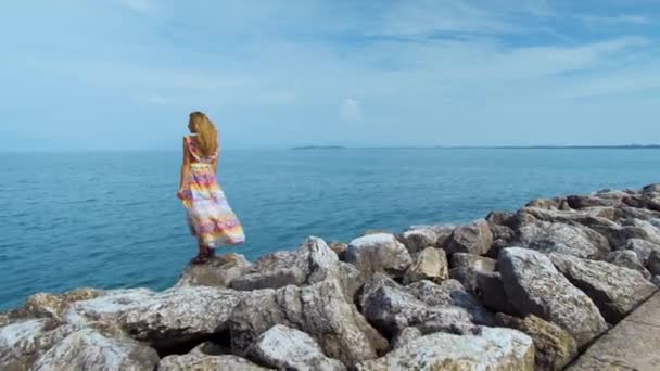 Chica en un hermoso vestido en piedras mirando al océano — Vídeo de stock