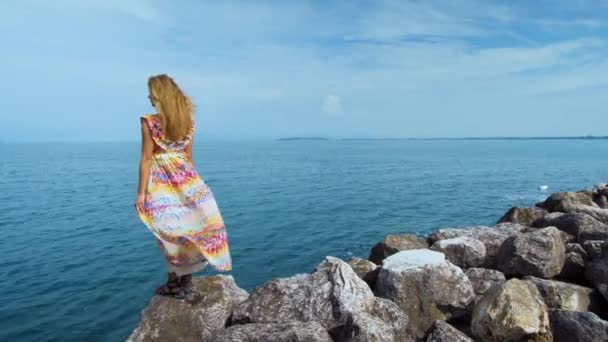 Девушка в красивом платье на камнях, смотрящая на океан — стоковое видео