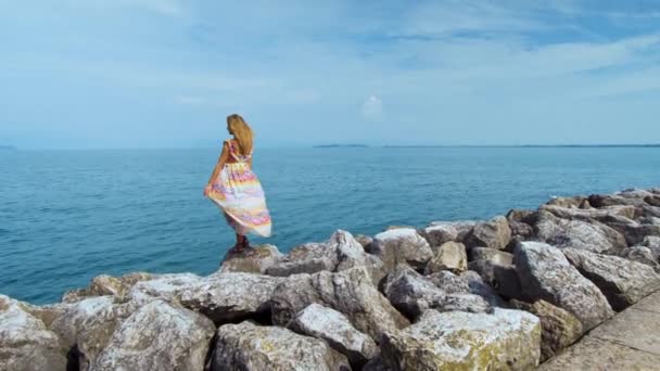 Девушка в красивом платье на камнях, смотрящая на океан — стоковое видео