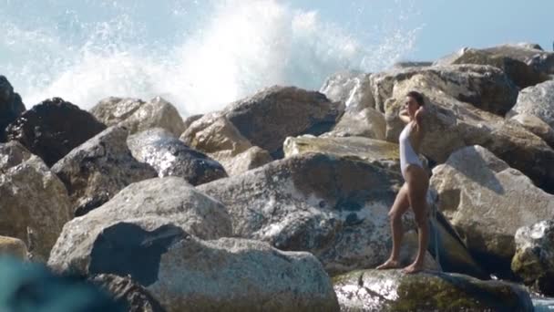 As ondas colidem contra as pedras, e enormes salpicos voam para a menina de pé em um maiô, câmera lenta — Vídeo de Stock