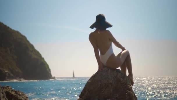Όμορφο κορίτσι σε ένα καπέλο κάθεται σε μια πέτρα σε ένα λευκό μαγιό και εξετάζει τη θάλασσα σε ένα γιοτ — Αρχείο Βίντεο