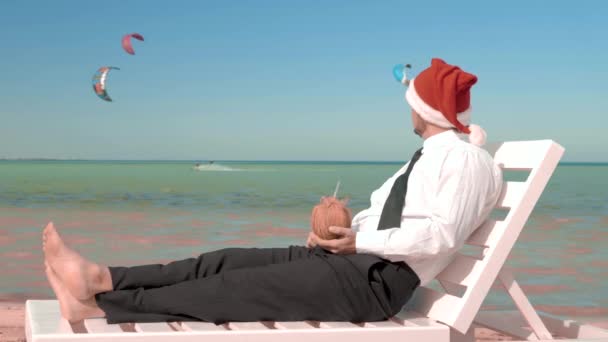 Businessman in santa claus hat sitting with a cocktail on the background of the sea — Stock Video