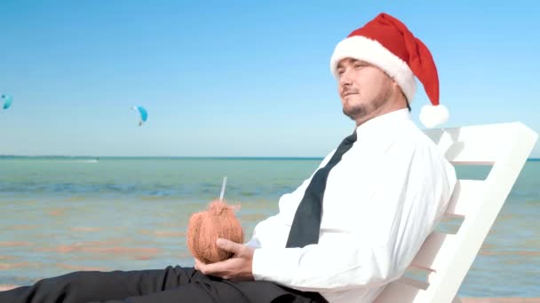 Бизнесмен в шляпе Санта-Клауса сидит с коктейлем на фоне моря — стоковое видео