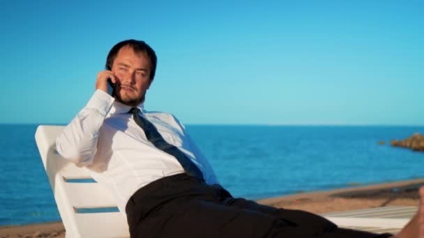 Молодой бизнесмен работает на пляже. Мужчина-профессионал сидит в удобном кресле — стоковое видео