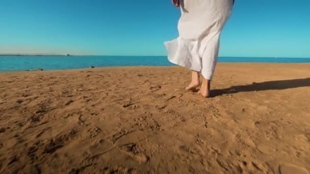 Legs of caucasian girl wearing white long dress walking barefoot sand on sea beach — Stock Video