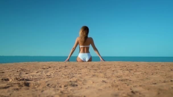 Das schöne Mädchen in einem weißen Badeanzug sitzt auf dem Hintergrund des Meeres — Stockvideo
