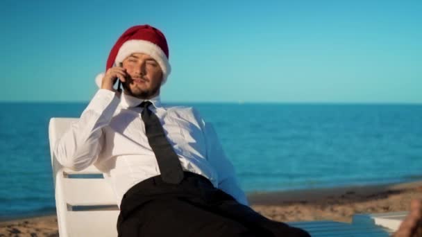 Podnikatel v santa claus klobouk sedí na lehátku na pozadí moře — Stock video