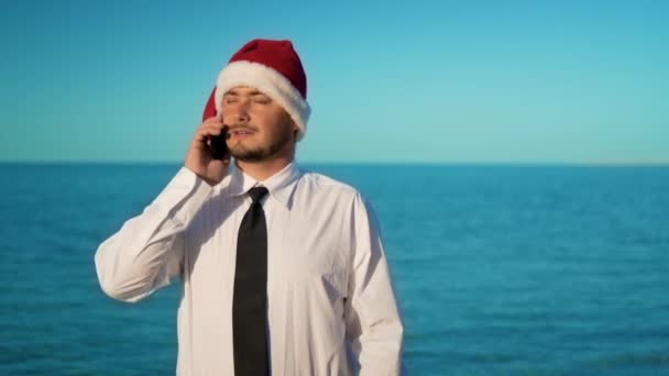 The businessman on the background of the beautiful sea talking on the phone, solving in cases in santa claus hat — Stock Video