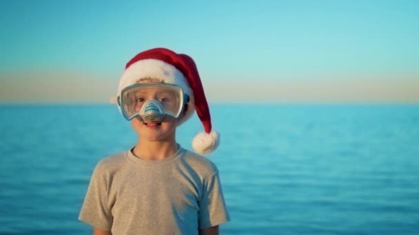 De jongen in de kerst hoed staat tegen de zee in duikbril en snorkel — Stockvideo