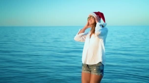 Bella ragazza che ascolta musica al telefono al mare con un cappello di Babbo Natale, indossa occhiali da sole, indossa un costume da bagno bianco e cuffie bianche, uno sfondo di acqua blu mare . — Video Stock