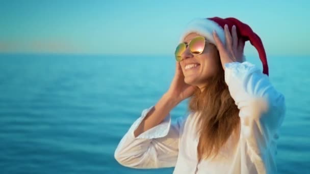 Menina bonita ouvindo música no telefone no mar em um chapéu de Papai Noel, usando óculos de sol, vestindo um maiô branco e fones de ouvido brancos, um fundo de água azul marinho . — Vídeo de Stock