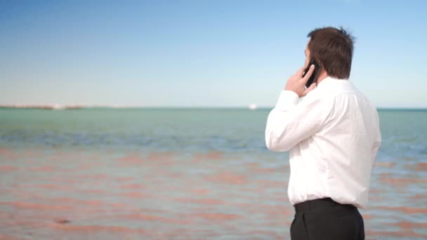 Joven hombre de negocios hablar en el teléfono celular cerca del mar — Vídeos de Stock