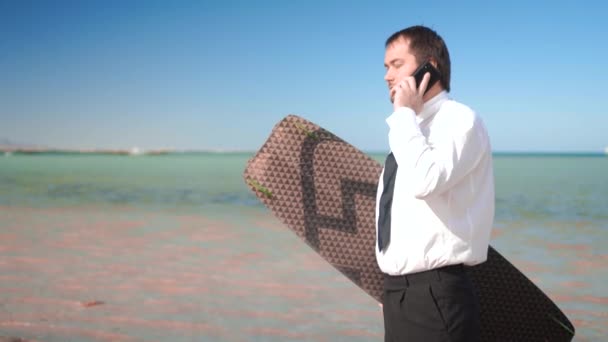 Podnikatel stojí na pobřeží a drží desku z kitesurfing — Stock video