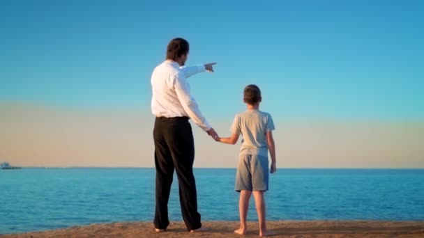 Il padre e il figlio in piedi, per mano, sulla riva del mare, il padre punta il dito in lontananza — Video Stock