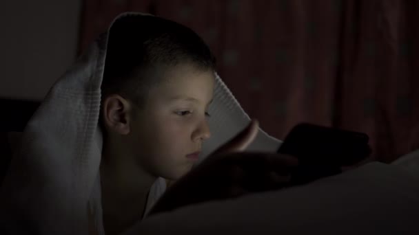 Un garçon heureux se trouve au lit sous une couverture et joue sur une tablette dans un jeu dans le noir. Le visage de l'enfant est éclairé par un moniteur lumineux — Video