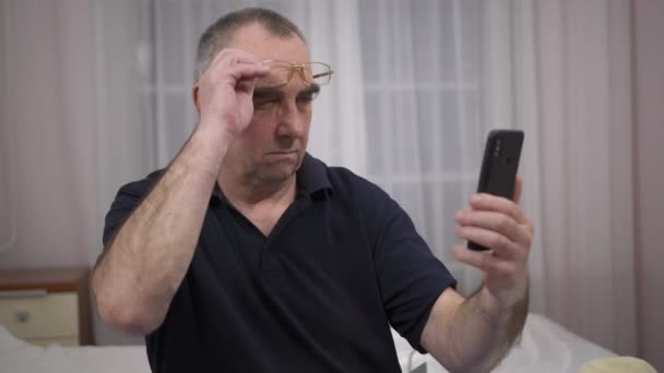 Пожилой человек в очках пытается что-то увидеть в телефоне — стоковое видео