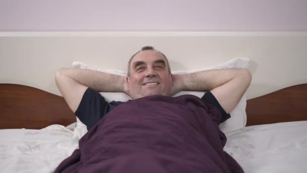 Un anciano yace en su cama descansando, sintiendo felicidad, alegría, consuelo — Vídeos de Stock