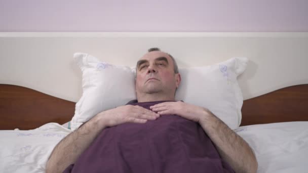Пожилой человек лежит в постели и отдыхает. Отгоняет мух и комаров, которые мешают ему отдыхать. — стоковое видео