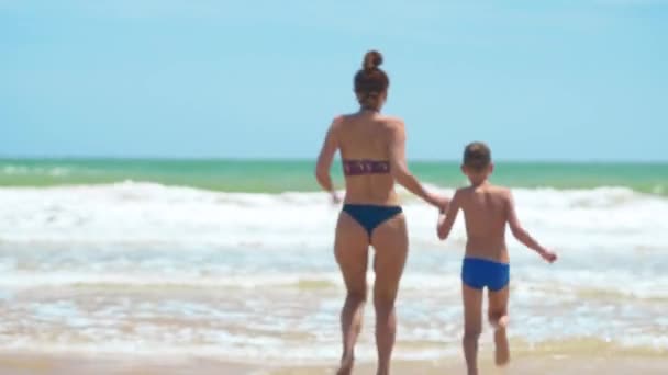 Feliz familia mamá y bebé corriendo descalzos sobre arena mojada, vista trasera. Mamá y bebé tomados de la mano juntos corriendo en la playa . — Vídeo de stock