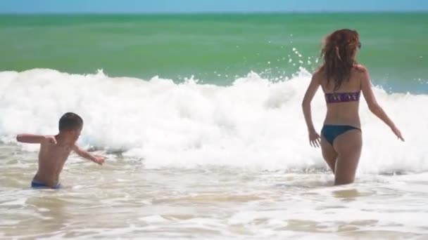 A criança bronzeada feliz feliz nadando e pulando com a mãe nas ondas do mar no dia ensolarado de verão na praia tropical . — Vídeo de Stock