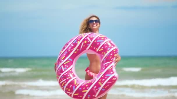 Mulher se divertindo e sorrir dançando com donut rosa. menina em bicini usando óculos de sol na praia. Mulher sexy bonita em férias de viagem de verão . — Vídeo de Stock