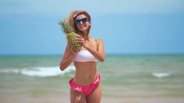 Menina sorrindo engraçado em trajes de banho de verão e óculos de sol. Mulher sexy com abacaxi fresco. Dança positiva perto do belo mar . — Vídeo de Stock