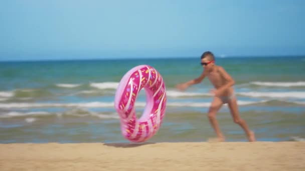少年はピンクの膨脹可能なドーナツとビーチに沿って実行し、海の背景に対して砂に沿ってそれを転がします。リラクゼーションと楽しみの概念. — ストック動画
