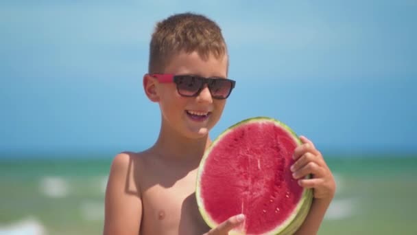 Pojken i glas håller en ljus vattenmelon på bakgrunden av havet. — Stockvideo