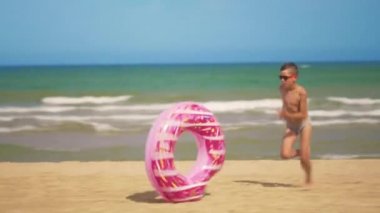 Çocuk pembe bir şişme çörek ile plaj boyunca çalışır, denizin arka planına karşı kum boyunca rulo. Rahatlama ve eğlence kavramı.