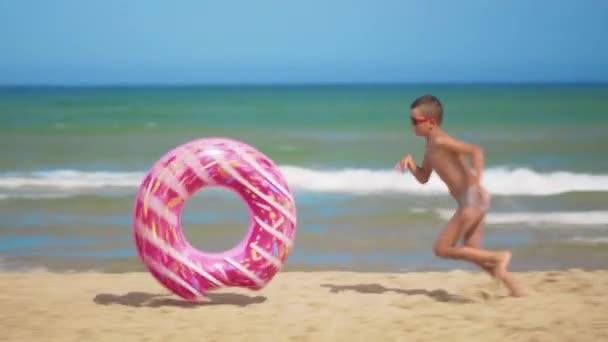 Το αγόρι τρέχει κατά μήκος της παραλίας με ένα ροζ φουσκωτό ντόνατ, το κυλάει κατά μήκος της άμμου στο φόντο της θάλασσας. Η έννοια της χαλάρωσης και της διασκέδασης. — Αρχείο Βίντεο