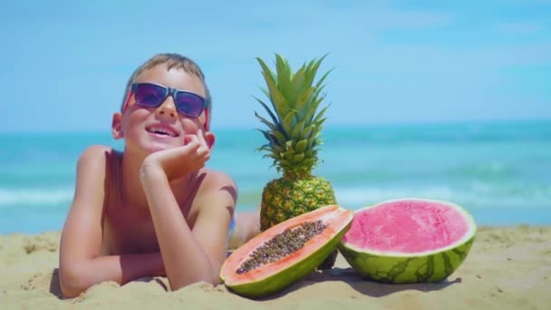 Niño sonriente se encuentra en la orilla del mar con una composición de frutas exóticas contra el mar . — Vídeos de Stock