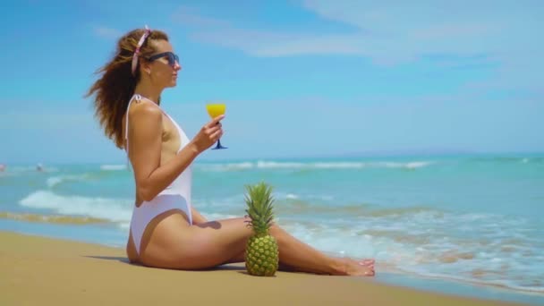 Fröhlich schöne junge Mädchen sitzen am Strand am Meer mit Ananas und Saft. — Stockvideo