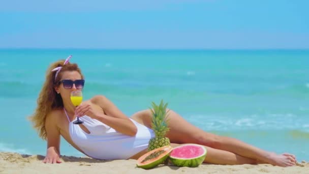 Die Frau trinkt hinter dem Meer frischen Orangensaft. junge schöne Frau am Meer auf dem Sand liegend, trinkt Orangensaft. Konzept Urlaub. — Stockvideo